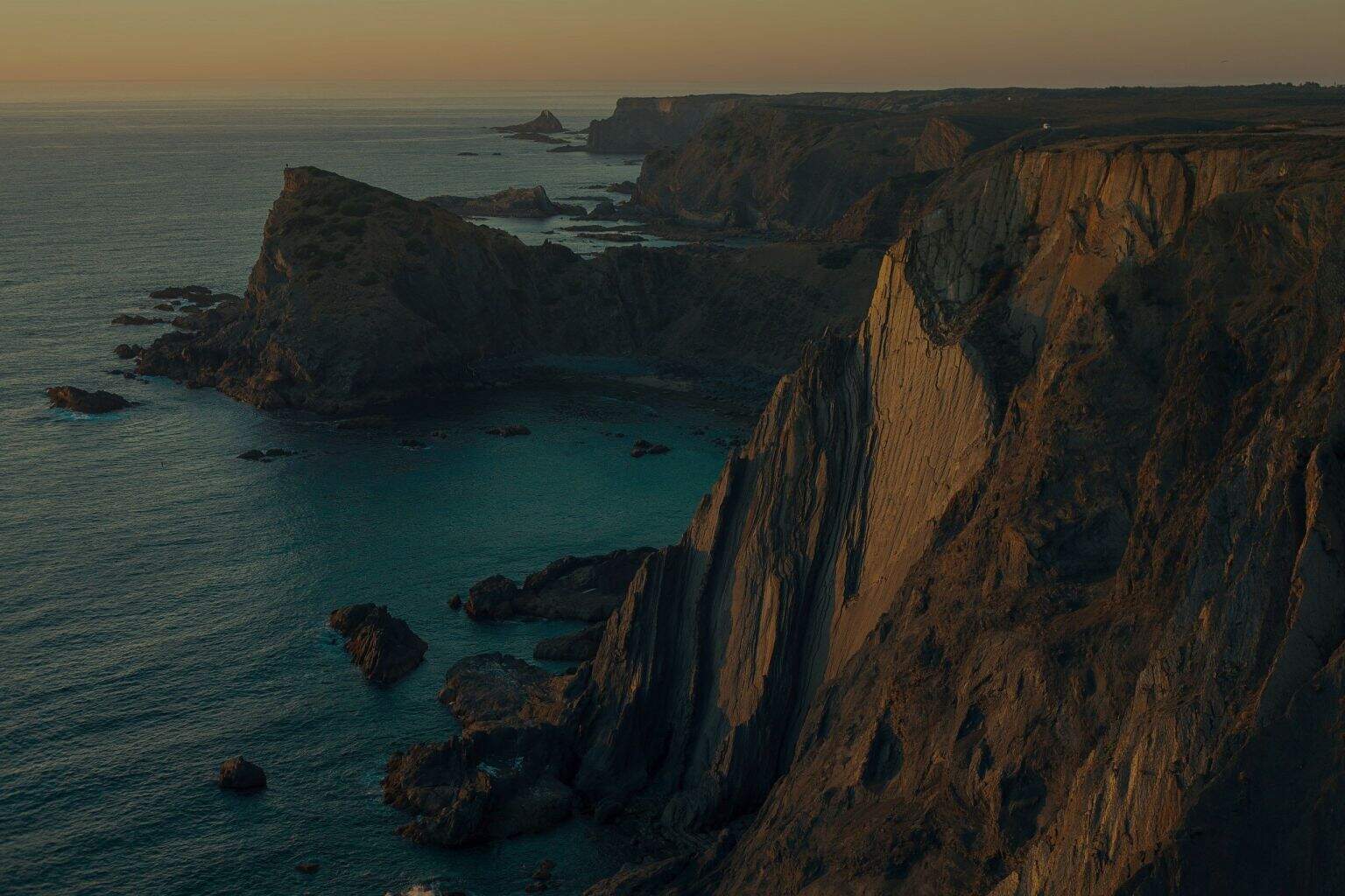 Vedere de pe coastă cu stânci înalte și valuri în ocean la apus.