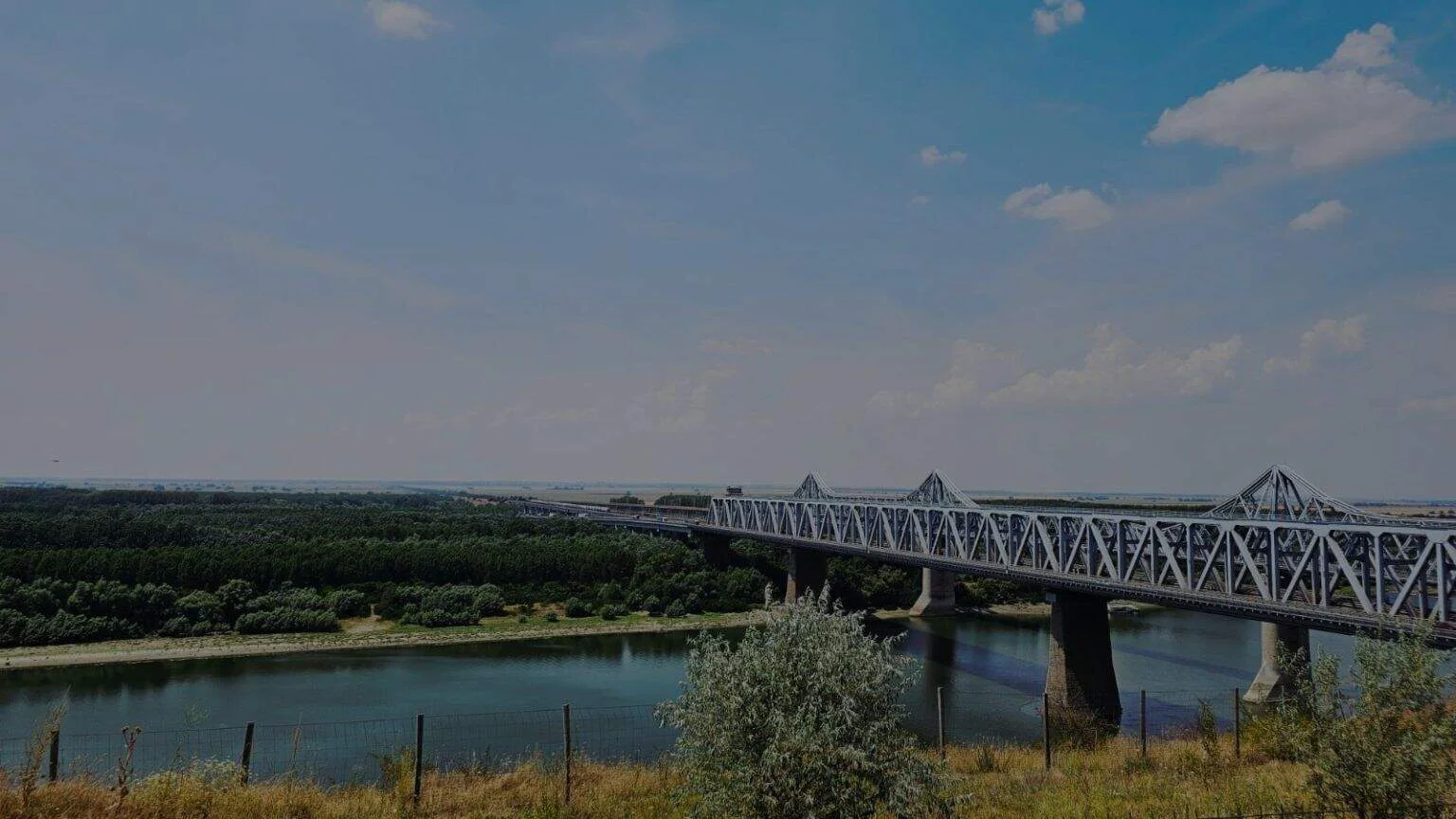 Pod peste râu, în peisaj natural, cu cer senin și vegetație verde.